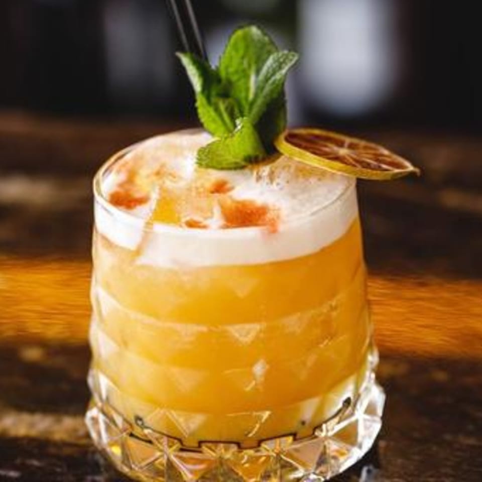 A vibrant orange cocktail with a foamy top, garnished with mint and a dried citrus slice.