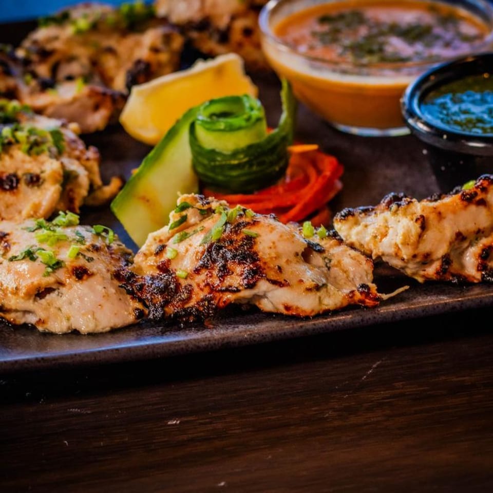 Grilled chicken tikka with lemon, cucumber, and two dipping sauces on a dark plate.